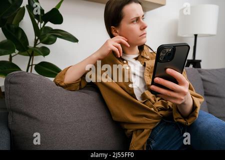 Fuoco selettivo di donna giovane pensierosa seduta sul divano, guardando da parte tenendo smartphone, in attesa di messaggio dall'uomo amato, sentendosi soli o. Foto Stock