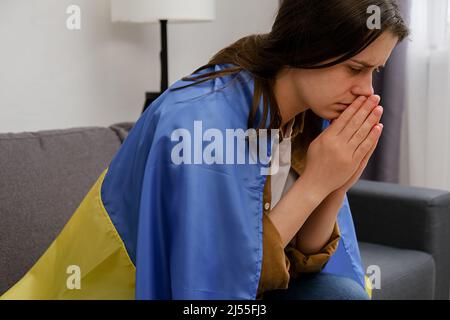 Vista laterale della triste giovane donna Ucraina con bandiera Ucraina seduta da sola sul divano a casa. Fermare la guerra in Ucraina. Ansia e anticipazione. Pace, Foto Stock