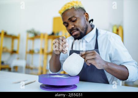 misto di razza afro maschio vasaio con grembiule nero seduto al vasaio da tavolo da officina Foto Stock