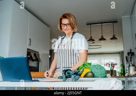 Donna di mezza età seguendo un tutorial di cucina video corso sul portatile mentre si prepara il pasto in una cucina. Donna che cucina sano piatto, pesce e verdure Foto Stock