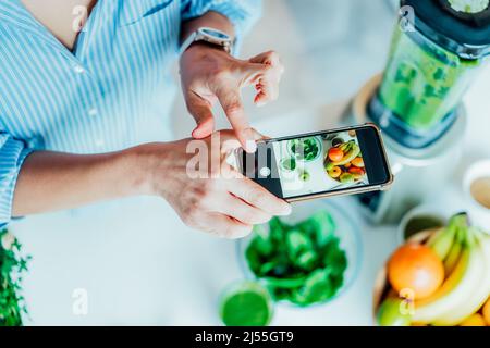 Blogger creazione di contenuti per i social media. Donna che fa foto, video con appena fatto vetro di disintossicazione verde frullato e si tratta di ingredienti in cucina Foto Stock