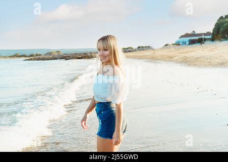 Donna sorridente felice in cima bianca e jeans pantaloncini godendo il momento durante una passeggiata sulla spiaggia di mare. Vacanze estive, vacanze. Piacere semplice. Foto Stock