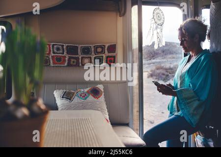 La donna adulta sorride e usa il telefono cellulare per inviare il messaggio in piedi sulla porta del suo camper casa alternativa. Turista che si gode di viaggio con roulotte Foto Stock