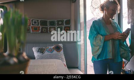 La donna adulta sorride e usa il telefono cellulare per inviare il messaggio in piedi sulla porta del suo camper casa alternativa. Turista che si gode di viaggio con roulotte Foto Stock