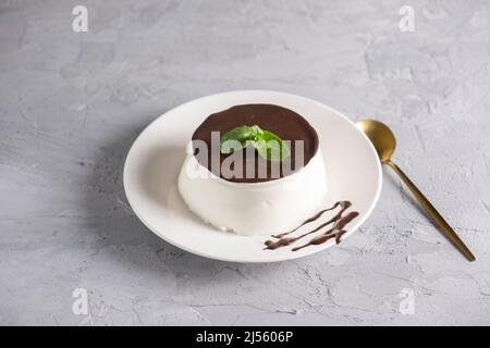 Dessert italiano panna cotta servita su piatto bianco con salsa di cioccolato e menta fresca su sfondo grigio chiaro Foto Stock