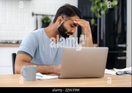 Fatica sovraccarica turbato indiano o arabo uomo, manager o freelancer, sedersi ad una scrivania in cucina con computer portatile, con gli occhi chiusi, in bisogno di riposo in una situazione stressante, hanno mal di testa, emicrania, Foto Stock