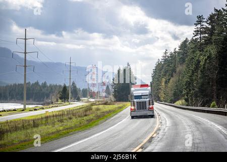 Standard industriale potente semirimorchio con carro grande e carico arancione classico con semirimorchio a pianale su strade strette con potenza ad alta tensione Foto Stock
