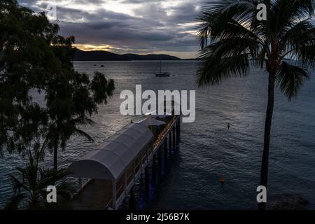 Un molo coperto di bianco in un resort turistico illuminato con luci fairy contro un cielo oscuro con yacht sull'oceano e alberi in primo piano. Foto Stock