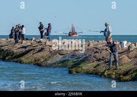 Pescatori sul molo del canale della nave Brazos Santiago Pass a South Padre Island, Texas. Una barca di gamberi esce dal canale come un pescatore lancia un Foto Stock