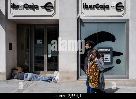 Un uomo senza tetto è visto dormire all'ingresso della società spagnola di servizi bancari finanziari Ibercaja come i pedoni passino oltre in Spagna. Foto Stock
