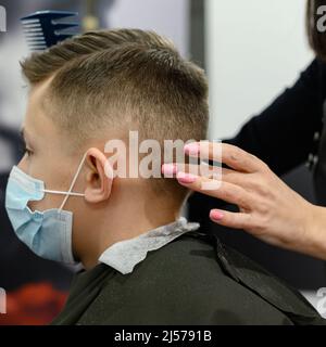 Un ragazzo adolescente ottiene un taglio di capelli in un barbiere durante una pandemia, un taglio di capelli nel salone, un cliente e un parrucchiere in maschere, un taglio di capelli del bambino con sciss Foto Stock