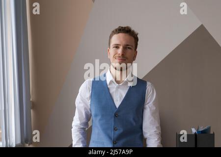 Concetto di fiducia e di business Ritratto giovane imprenditore di successo in maglia blu bianco gilet sorridente ampiamente con espressione sicura di sé con barba guardando la fotocamera contro parete di fondo in ufficio Foto Stock