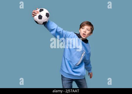 Gioioso giovane uomo in felpa blu che posa isolato su sfondo blu parete. Persone sincero emozioni stile di vita concetto. Spazio di copia del layout. Tenere la palla di calcio, fare il gesto del vincitore, yelling adolescente Foto Stock