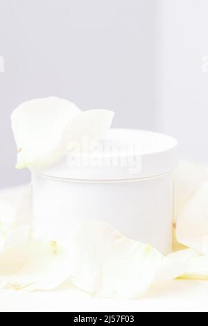 Vaso bianco con crema tra delicati petali di fiori di rosa bianca su sfondo chiaro. Cosmetici naturali biologici concetto, primo piano, mock-up Foto Stock
