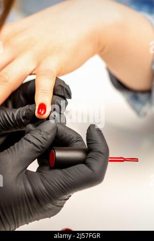 Vernice per manicure. Primo piano di un maestro manicure che indossa guanti di gomma nera applicando vernice rossa su un'unghia femminile nel salone di bellezza Foto Stock