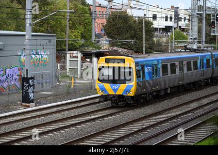 Il treno Comeng, che presenta l'attuale livrea PTV, passa attraverso uno dei quartieri dello shopping metropolitani di Melbourne, con negozi e traffico in background Foto Stock