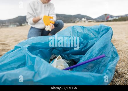 Una donna in guanti con una speciale borsa blu raccoglie rifiuti tra la sabbia lungo la costa. Il problema dell'inquinamento ambientale. Pulizia dei rifiuti Foto Stock