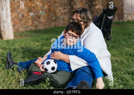 Una mamma che gioca con suo figlio che ha una disabilità che tiene una palla di calcio nel parco Foto Stock