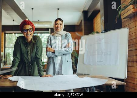 I designer musulmani felici sorridono alla macchina fotografica mentre lavorano sui disegni del progetto. Due donne d'affari creative che progettano un nuovo progetto in un ufficio. Arco Foto Stock