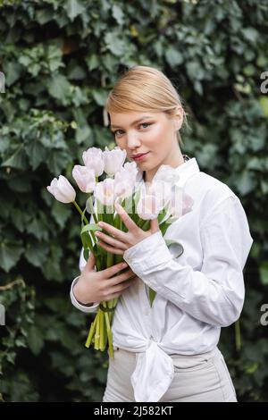 donna bionda in camicia bianca che tiene tulipani bianchi e guarda la macchina fotografica vicino all'edera verde Foto Stock