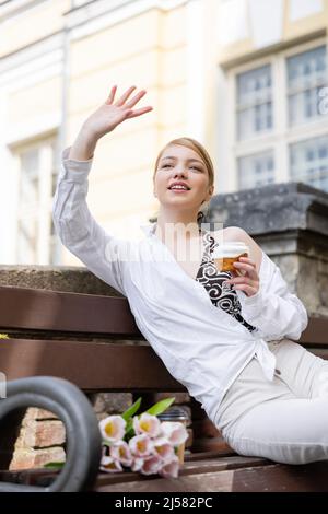 donna sorridente con cupcake che agita la mano mentre si siede sulla panca Foto Stock