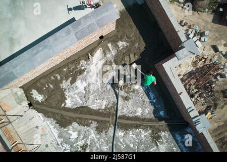 Vista aerea di una pala per muratore di sabbia-cemento mentre si lavora all'aperto. Operatore maschile che utilizza una macchina per la miscelazione di rasatori per calcestruzzo durante la preparazione del materiale del rasatore per pavimenti sul tetto del nuovo edificio. Foto Stock