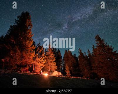 Campeggio notturno sulla collina di montagna. Vista dal basso di due campeggiatori seduti l'uno di fronte all'altro vicino al fuoco vicino alla tenda turistica. Coppia di amici che si riuniscono vicino falò in una calda serata stellata. Foto Stock