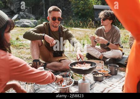 Sorridendo giovane uomo caucasico in occhiali da sole seduti tra amici e frittura uova sul bruciatore in campeggio Foto Stock