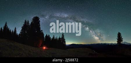 Vista panoramica delle colline di montagna serali sotto il cielo stellato. Fiammella piccola, silhouette di tenda vicino agli alberi sullo sfondo di favoloso paesaggio di Via Lattea galassia e montagne innevate. Foto Stock