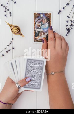 Le mani con chiodi viola e anelli tengono il mazzo di carte Tarocchi su una superficie bianca con lanterna e fiori di lavanda. Aprire la scheda con l'iscrizione Vasilisa Foto Stock