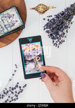 Le mani con chiodi viola e anelli tengono Tarot card su una superficie bianca con lanterna e fiori di lavanda. Vista dall'alto. Minsk, Bielorussia - 28.07.2021 Foto Stock