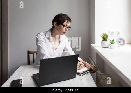 Donna di mezza età con bruna dai capelli corti in occhiali lavora su un computer portatile vicino alla finestra. Scrive con attenzione in un notebook Foto Stock