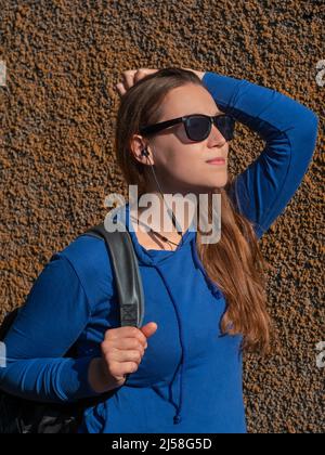 Giovane donna con cappuccio blu e occhiali da sole neri su sfondo intonaco per ascoltare musica e podcast con cuffie Wi-Fi. Signora hipster in wirel Foto Stock