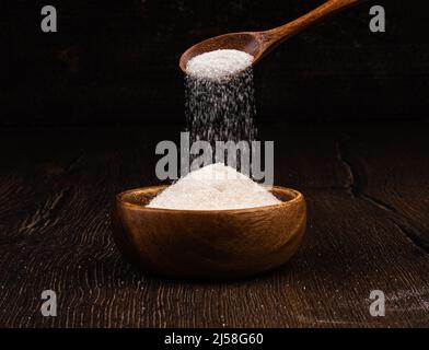 Lo zucchero granulato bianco è versato da un cucchiaio di legno in una ciotola di legno. Spazio di copia. Foto Stock