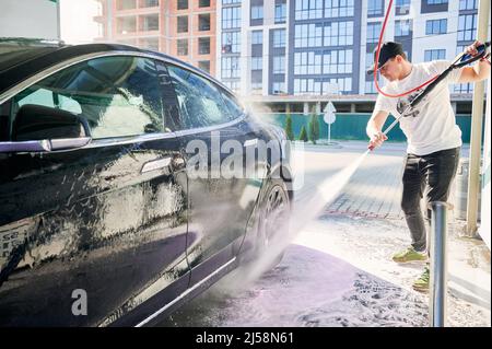 Maschio pulitore lavando le porte laterali di costoso automobile nera con acqua pressurizzata. Pulizia automobile con acqua dalla pistola. Potente spruzzatore sciacquare le finestre laterali sopra le porte di auto. Foto Stock