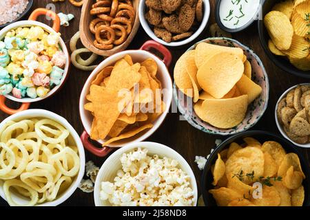 Cibo o spuntini malsani. Tutti i classici spuntini di patate con arachidi, anelli di popcorn e cipolle e pretzel salati in piatti da ciotola su vecchio sfondo di legno. Foto Stock