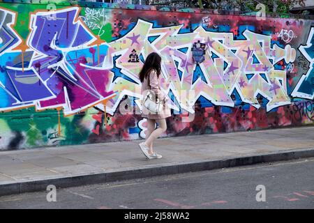 Giovane donna che indossa rosa a piedi da colorati graffiti pittura hoardings a Brick Lane Shoreditch primavera Aprile 2022 Londra Inghilterra Regno Unito KATHY DEWITT Foto Stock