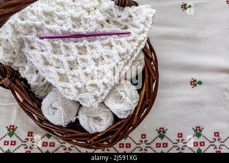 Coperta parzialmente rifinita con un motivo originale a punto di crochet in rilievo, uncinetto viola e batuffoli di cotone in un cestino di vimini marrone, tovaglia Foto Stock