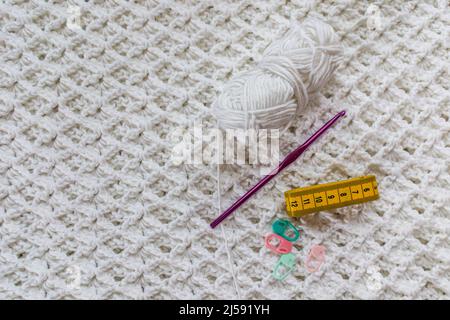 Vista dall'alto di una coperta bianca con un motivo a punto a crochet in rilievo, un batuffolo di cotone, pennarelli verdi e rosa, metro a nastro giallo e viola metallico c Foto Stock