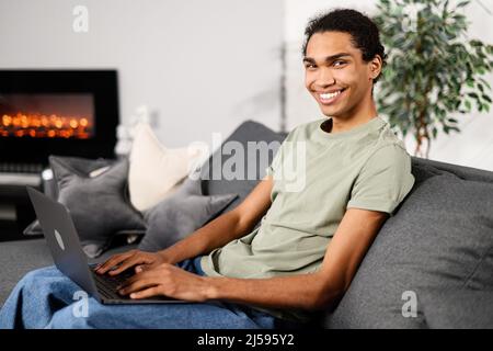 Vista laterale dell'allegro uomo multirazziale indossando abbigliamento casual seduto sul divano e utilizzando un computer portatile in un moderno ufficio domestico. Felice imprenditore maschio digitando sulla tastiera e sorridendo alla fotocamera Foto Stock