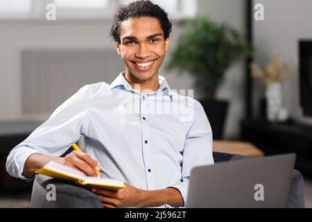 Occupato ragazzo freelance multietnico che utilizza un computer portatile per lavoro o per studiare a distanza da casa. Uomo concentrato che guarda il webinar online e prende appunti in notebook, e-learning Foto Stock