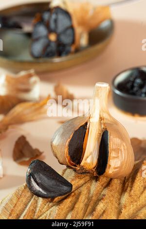 Testa fermentata d'aglio e spicchio d'aglio su un piatto di legno fatto a mano. Una piccola ciotola di argilla con pasta d'aglio nera sullo sfondo. Sano e fermentato Foto Stock