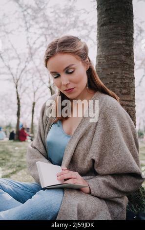 Bella giovane donna che ha pic-nic e libro di lettura sul giorno di primavera soleggiato nel parco durante la stagione di fioritura dei ciliegi. Festa della fioritura dei ciliegi. Ricrea all'aperto Foto Stock