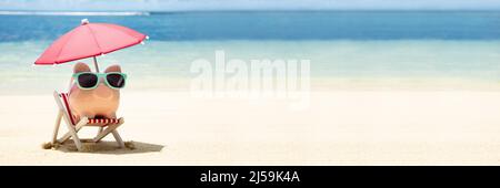 Rosa Piggybank con occhiali da sole e ombrellone sulla sedia a sdraio sulla sabbia in spiaggia Foto Stock