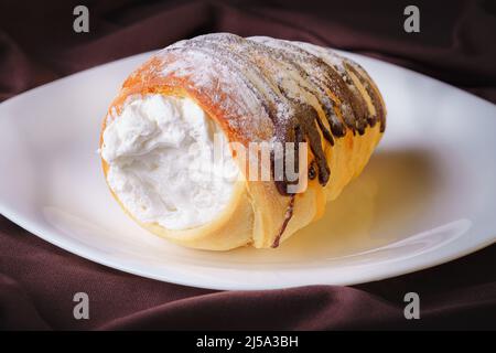 delizioso pane arrotolato a forma di cono ripieno di crema di pasticceria ricoperta di cioccolato fatto in casa e zucchero posto su un piatto di ceramica bianca Foto Stock