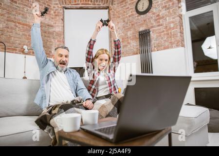 Famiglia caucasica emozionale seduta sul divano con le mani in su mentre festeggia la vittoria durante i video giochi sul laptop. Coppia matura seduta sul divano e godersi il tempo libero con divertimento. Foto Stock