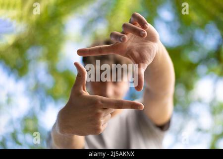 Un bel giovane uomo gli fa lampeggiare gli occhi e guarda attraverso una cornice fatta di mani. L'uomo attraente fa cornice dalle dita, isolato su sfondo sfocato wit Foto Stock