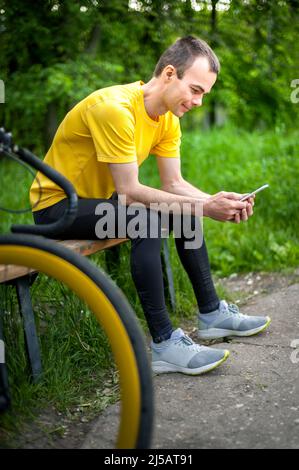 Un giovane seduto su una panchina in un parco pubblico comunica tramite comunicazione mobile. Sullo sfondo con la sua moto. Tra alberi e vegetazione. Foto Stock
