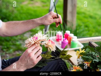 Arte e mestieri adulti concetto di creatività terapia d'arte, l'uomo dipinge fiori di seta in ambiente tranquillo all'aperto con sfondo verde erba e stampelle appoggiate Foto Stock