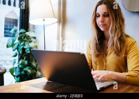 bella giovane donna che indossa una camicia gialla che digita sul suo laptop seduto in un bar Foto Stock